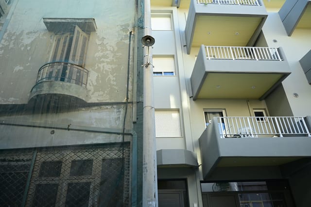 Overgrown courtyard visible through a rusted gate opposite renovated apartments